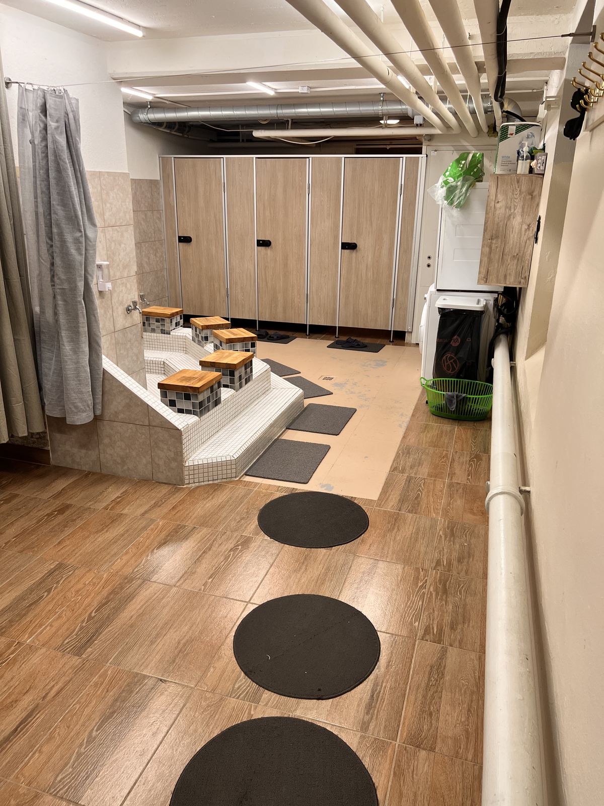 Waschbereich mit gefliester Bank, hölzernen Sitzhockern über den Becken und Kabinentüren aus Holz.